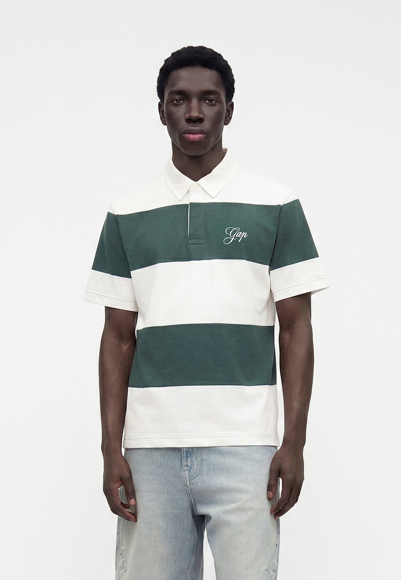Jeune homme portant un polo rayé vert et blanc avec le logo "Gap" et un jean bleu clair, debout devant un fond blanc uni.