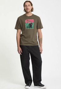 T-shirt di cotone verde oliva con un disegno grafico colorato raffigurante una figura e dei fiori. Abbinata a pantaloni neri larghi e sneakers nere.