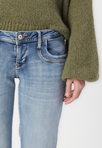 Hellblaue Jeans mit leichtem Used-Look, ausgestattet mit einer Vorderstasche und einem Knopfverschluss. Kombiniert mit einem strukturierten, olivgrünen Strickpullover.