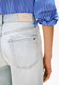 Svetlomodré denimové džínsy majú na zadnom vrecku malú kontrastnú značkovú nálepku. Pás je ozdobený béžovou etiketou s nápisom "TOMMY HILFIGER."
