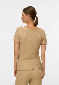 Donna con capelli castani legati in una coda di cavallo bassa, che indossa un top a maniche corte beige testurizzato e pantaloni abbinati, vista da dietro su uno sfondo bianco.