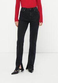 Black denim jeans with a high waist, straight leg design, and side slits. Paired with a red long-sleeve top and black heeled shoes.