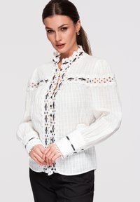 Femme portant un chemisier blanc avec bordure en dentelle et accents en ruban noir, debout devant un fond clair uni, les mains doucement jointes.