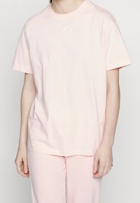 Světle růžové bavlněné tričko s krátkým rukávem, kulatým výstřihem a malým bílým logem Nike na hrudi. V kombinaci se sladěnými růžovými kalhotami.