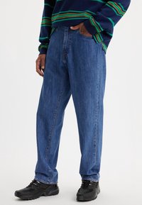 Modré džíny s volným střihem, se viditelným šitím a vysokým pasem. V kombinaci s černými sportovními botami.