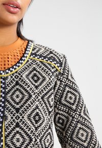 Veste à motifs avec un design géométrique noir et blanc agrémenté d'accents jaunes et noirs. Le matériau semble tissé avec des détails texturés.