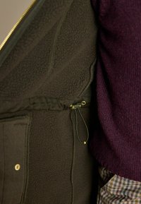 Tmavohnedrá fleece bundy s vnútorným zipsovým vreckom a nastaviteľnou šnúrkou. Svetlofialový pletený sveter je viditeľný v blízkosti.