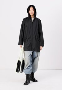 Manteau noir imperméable avec capuche, fermeture éclair et poches latérales. Porté avec un jean large bleu clair et des bottines noires, tenant un sac.