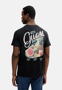 Camiseta de algodón negra con un gráfico colorido de frutas y la palabra "Guess" en cursiva en la parte posterior. Diseño de manga corta y cuello redondo.