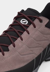 Κοντινό πλάνο ενός αθλητικού παπουτσιού με επάνω μέρος από μαύρο και μοβ σουέντ, μαύρη γλώσσα με λευκό κυκλικό λογότυπο και κορδόνια μπορντό με μοτίβο.