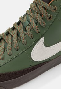 Zapatilla verde de cuero con un logo de ante gris texturizado, suela marrón oscuro y cordones verdes cosidos en naranja. Presenta un diseño clásico de caña alta.
