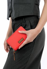 Leuchtend rotes Portemonnaie mit glatter, matter Oberfläche, versehen mit einem Akzent aus braunem Leder und einem Reißverschluss-Anhänger, gehalten in einer Hand vor einem grauen Outfit.