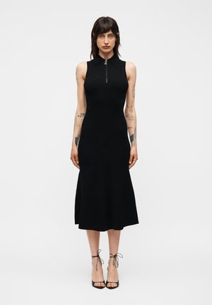 Kobieta z tatuażami nosi czarną, bezrękawnikową sukienkę midi z kołnierzykiem i zamkiem z przodu, połączoną z czarnymi sandałami na wysokim obcasie z paskami na białym tle.