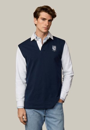 Junger Mann, der ein marineblaues ärmelloses Rugbyshirt über einem weißen Langarm-Poloshirt und hellblauen Jeans trägt, steht mit der Hand in der Tasche.