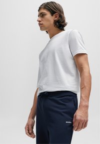 Weißes T-Shirt mit kurzen Ärmeln und V-Ausschnitt, kombiniert mit dunkelblauen Jogginghosen, die ein weißes Logo auf dem Oberschenkel haben. Glatter Stoff, lockere Passform.