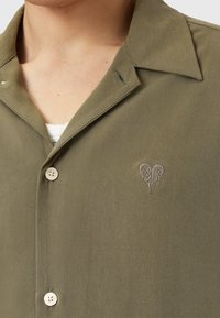 Olijfgroene blouse met een kraag, crèmekleurige knopen en een hartvormig geborduurd detail. De stof heeft een zachte textuur.