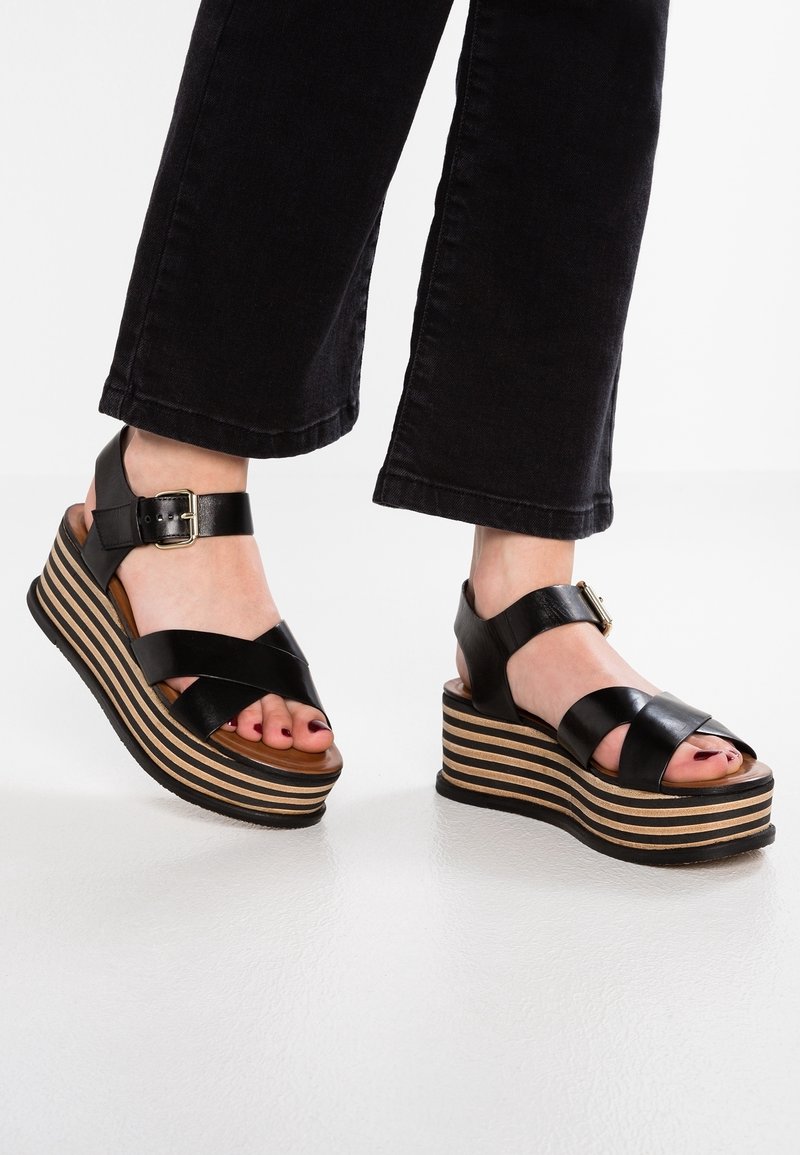 Black leather wedge sandals with crisscross straps, adjustable ankle buckle, and a striped wooden platform sole.