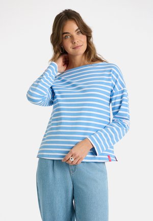 Junge Frau, die ein blau-weiß gestreiftes Langarmshirt und hellblaue Jeans trägt, den Hals berührt und den Saum des Shirts hält.