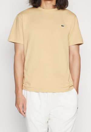 Person iført en beige kortærmet t-shirt med lille grønt logo på brystet og hvide bukser, stående mod en ensfarvet baggrund.