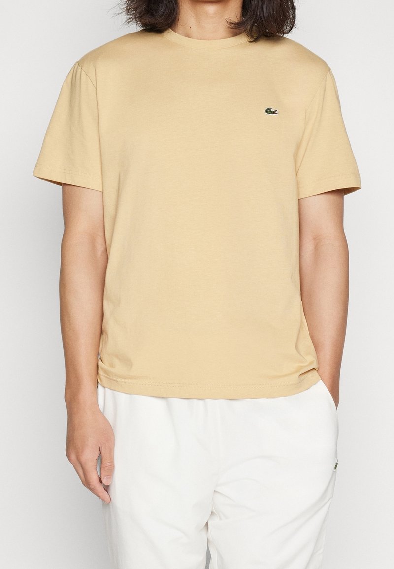 Personne portant un t-shirt beige à manches courtes avec un petit logo vert sur la poitrine et un pantalon blanc, debout contre un fond uni.