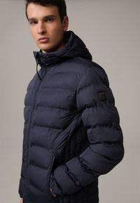 Navy puffer jacket with a hood, featuring quilted texture, zippered pockets, and a branded patch on the left sleeve.