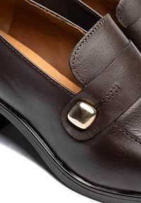 Braune Leder-Loafers mit abgerundetem Zehenbereich, glatter Textur und einem dekorativen Metallakzent auf dem Spann zur Verzierung. Entlang der Kanten ist eine sichtbare Naht zu erkennen.