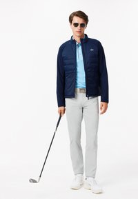 Giacca imbottita blu navy, polo azzurra, pantaloni grigi e scarpe da golf bianche. Tiene un bastone da golf e indossa occhiali da sole. Estetica sportiva pulita e moderna.