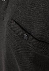Dunkelgrauer Stoff mit texturiertem Gewebe. Verfügt über eine Brusttasche mit einem schwarzen Knopf und einem dezenten Kragenabschluss.