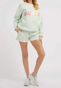 Jasnozielony sweatshirt z logo "GUESS" w kolorowej czcionce, w zestawieniu z pasującymi szortami, noszony z białymi i miętowymi trampkami. Casulowy, sportowy wygląd.