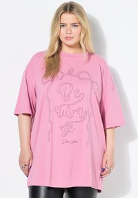 Rosa, oversized T-Shirt mit kurzen Ärmeln, das den Text "Be Aware" in schwarzer Umrandung zeigt, aus weichem Baumwollstoff mit glatter Textur.