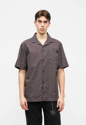 Joven con camisa marrón a cuadros de manga corta y pantalones negros con un diseño de estrella punteada en el muslo izquierdo.