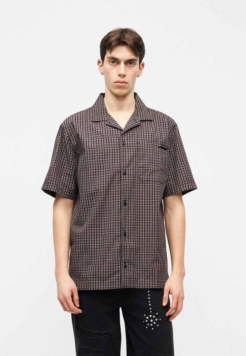 Νεαρός άνδρας που φοράει καφέ καρό πουκάμισο με κοντά μανίκια και κουμπιά, και μαύρο παντελόνι με διακοσμητικό σχέδιο από κουκκίδες σε σχήμα αστέρα στο αριστερό μηρό.