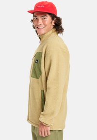 Beige fleece jacket featuring a green patch pocket, high collar, and zipper detail. Worn with a red cap. Soft texture and relaxed fit.