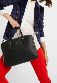 Bolso de mano de cuero negro con acabado texturizado, cierre de cremallera, dos asas superiores y una correa para el hombro desmontable. Accesorios de hardware en plata visibles.