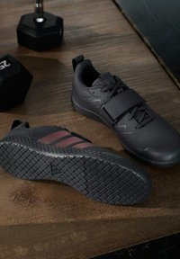 Para czarnych butów do podnoszenia ciężarów z teksturowaną podeszwą i paskiem, ustawiona na drewnianej podłodze obok dwóch sześciokątnych hantli o wadze 25 funtów.