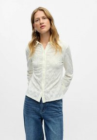 Donna con lunghi capelli biondi, che indossa una blusa bianca a righe con bottoni e jeans blu, in piedi contro uno sfondo chiaro e semplice.