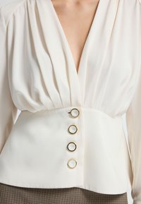 Cremefarbige Bluse mit tiefem V-Ausschnitt, gepufften Ärmeln, taillierter Passform und vier goldenen Akzentknöpfen auf der Vorderseite.