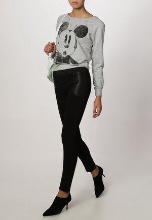 Kobieta ubrana w szarą bluzę z dużą czarno-białą kreskówkową twarzą, czarne dopasowane spodnie, czarne buty na obcasie, trzymająca małą białą torebkę.