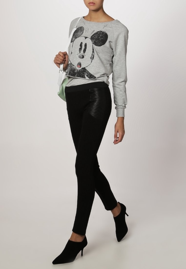Femme portant un sweat-shirt gris avec un grand visage de dessin animé noir et blanc, un pantalon noir ajusté, des chaussures à talons noires, et tenant un petit sac à main blanc.