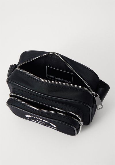 Sac bandoulière en nylon noir avec des zips en argent, deux poches externes et une étiquette de marque à l'intérieur. Texture douce et design épuré.
