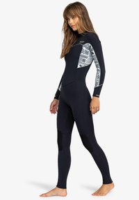 Traje de neopreno negro de manga larga, diseñado con patrones geométricos blancos en los hombros. Presenta paneles en las rodillas y una silueta ajustada.