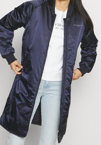 Marineblå lang pufferjakke, skinnende nylonmateriale, ribbede manchetter, lynlåslukning, sidelommer, båret over en hvid sweatshirt og lyse denimjeans.