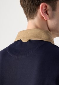 Maglione blu navy con colletto in corduroy color tan. Texture liscia, cuciture visibili e un dettaglio con anello sul retro accentuano il design.