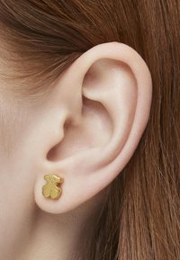 Złoty kolczyk w kształcie misia, eksponowany na uchu modelki, o gładkiej teksturze i zaokrąglonych krawędziach.