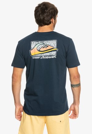 Homme aux cheveux foncés portant un t-shirt bleu marine avec un motif coloré de vague et de montagne et un short beige, dos à la caméra.