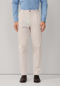 Muškarac nosi svijetlosmeđe krojene hlače, plavu košulju s dugim rukavima i tamnosmeđe kožne mokasine, stojeći protiv jednostavne pozadine.