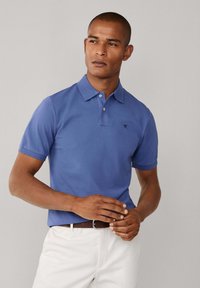 Blauwe poloshirt met een kraag en korte mouwen, met een klein zwart logo op de borst. Draagt een witte broek en een bruine riem.