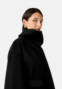 Cappotto nero in lana con colletto alto, tessuto strutturato, design oversize e vita aderente, abbinato a orecchini statement dorati.