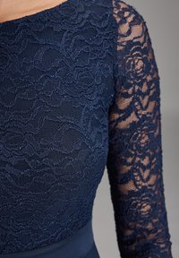 Marinblå spetstyg med blommönster täcker överkroppen och halvgenomskinlig ärm på en figurnära klänning.