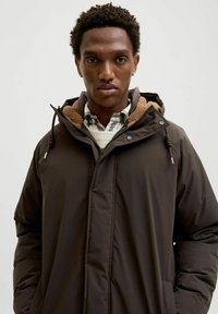 Brūna polsterēta jaka ar rāvējslēdzēja aizdari un regulējamu kapuci, ar mīkstu fliša odere. Pārsieta pāri raibam kreklam.